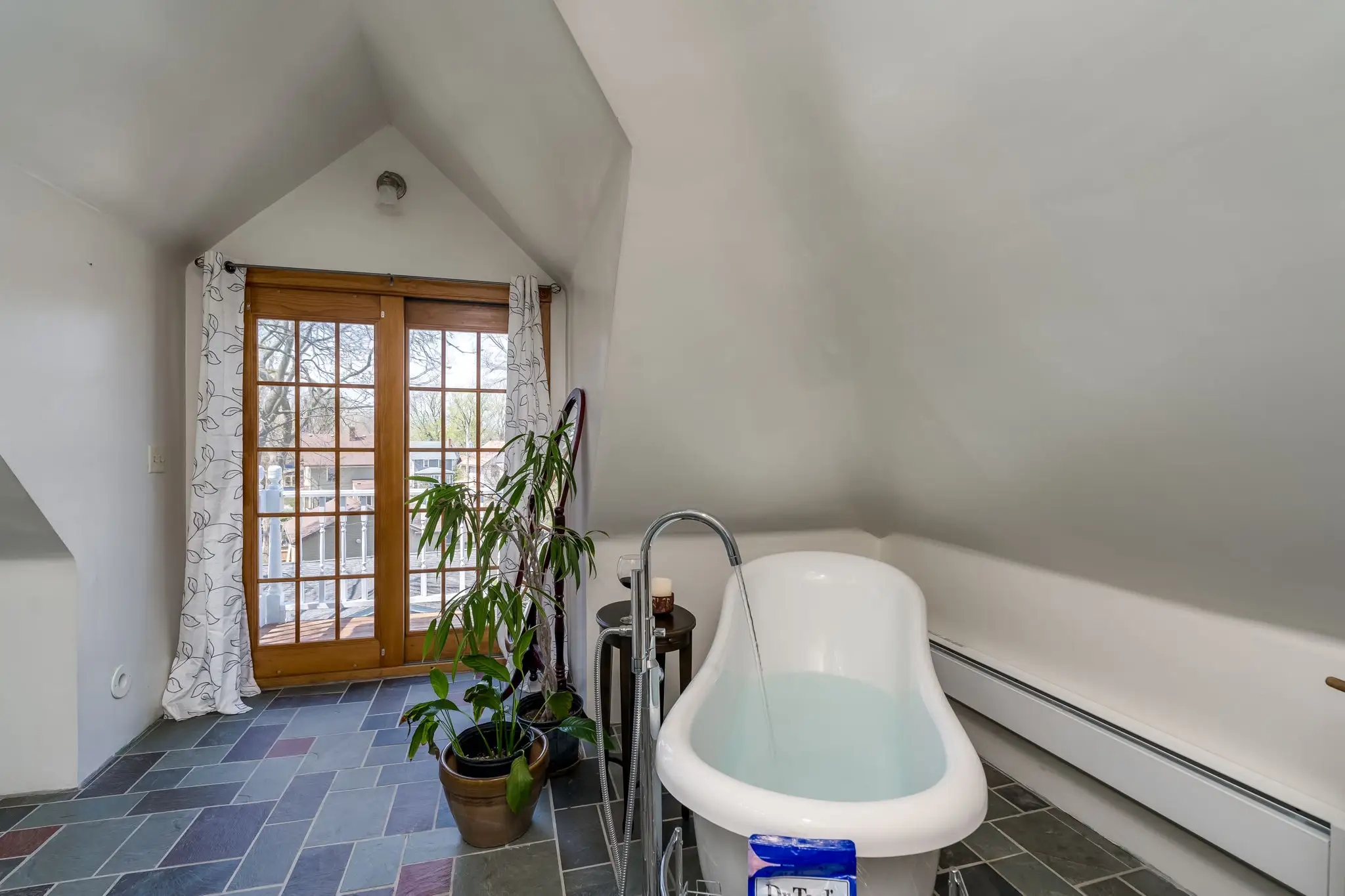 A bathroom with a large clawfoot tub with water pouring into it, a side table with a candle and glass of wine, a potted plant, and french doors leading to a balcony.