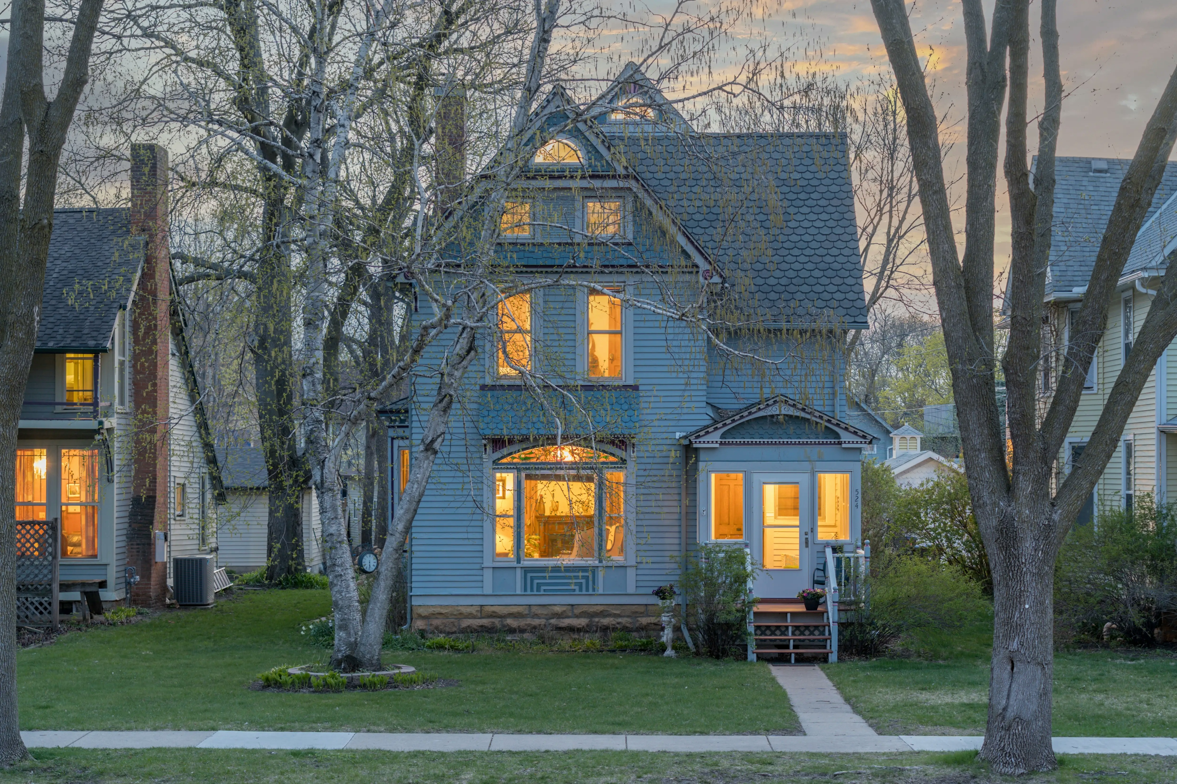 A 3 story blue Victorian house lit up at night, with green grass on the lawn and a mature tree in the middle of the front yard.