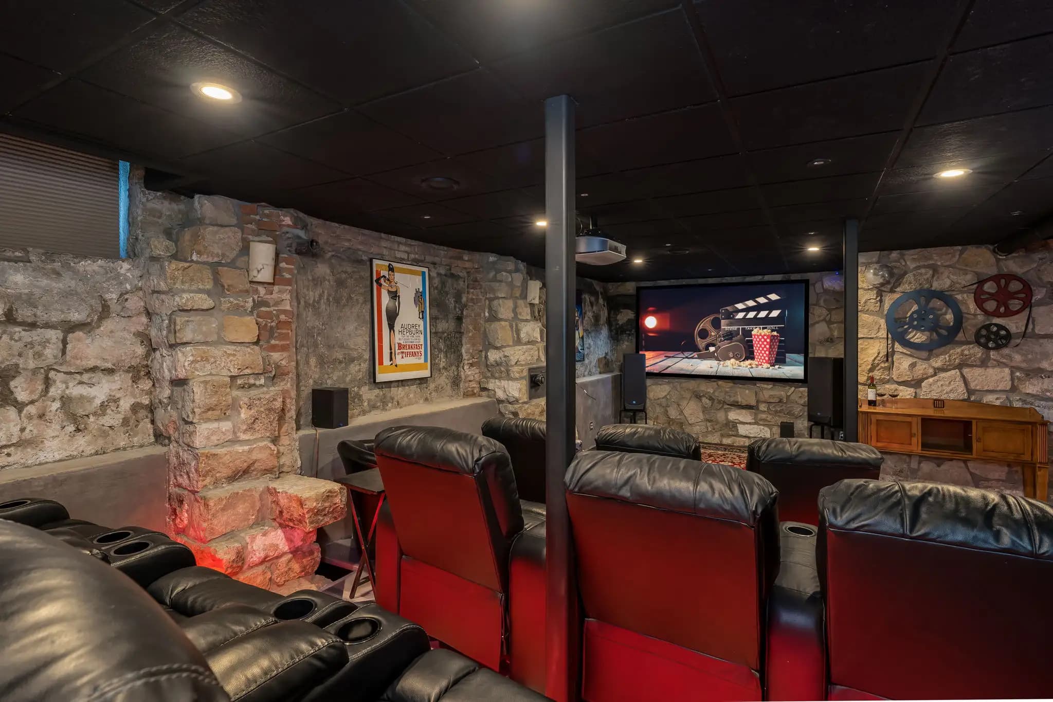 A cozy movie theater room with stone walls, black ceiling, red and black reclining chairs, a large TV at the front of the room, with movie posters and film, reels on the walls