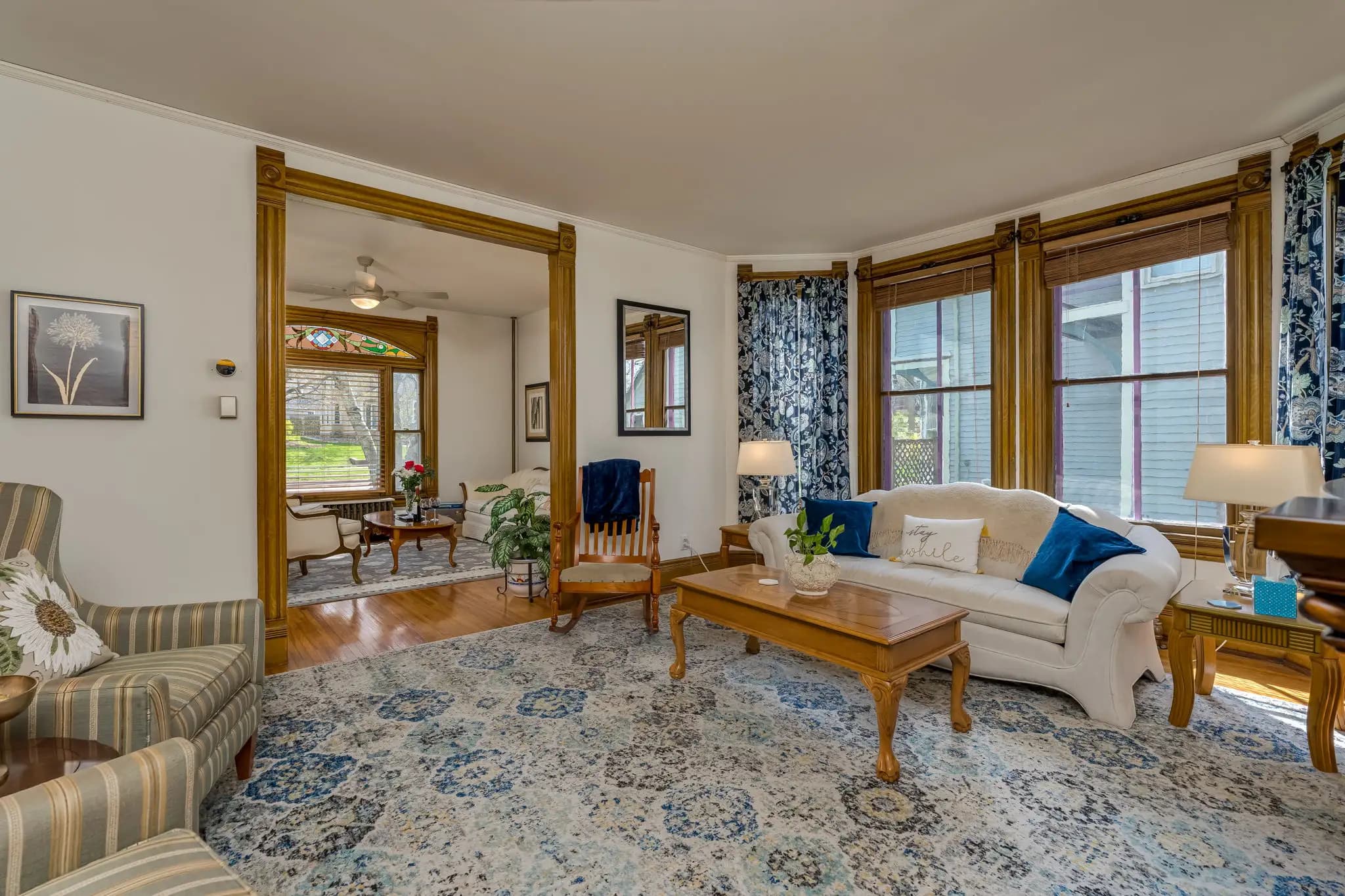 A spacious living room with a couch, stuffed armchairs, wooden coffee table and side chairs, and a rocking chair. A large open door leads to a parlor with more sitting areas.