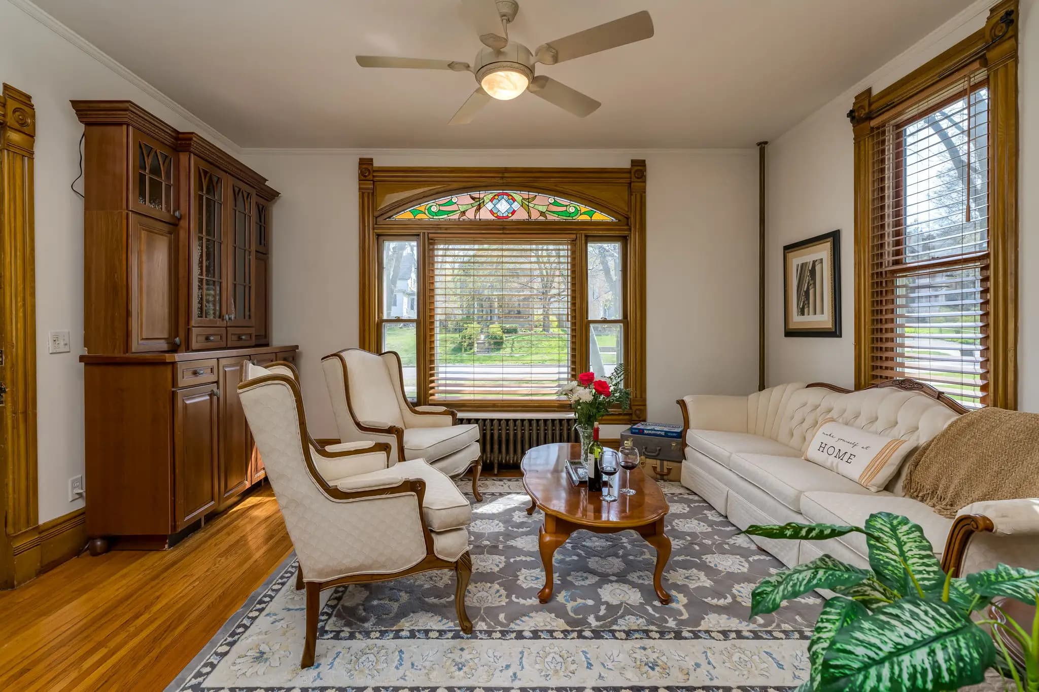 A cozy room with hardwood floors, Victorian style furniture, a wood coffee table, large china hutch, and a large window facing outside the front of the house with a beautiful stained glass on the top.