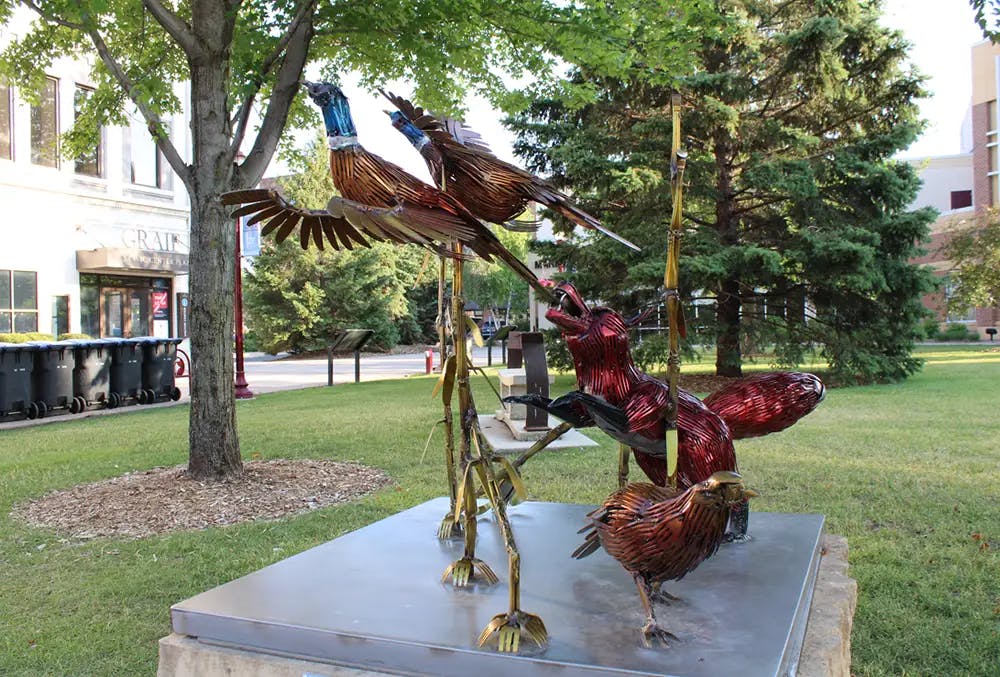 metal bird sculptures on a raised cement podium with a stainless steel top, in a city park with trees and green grass