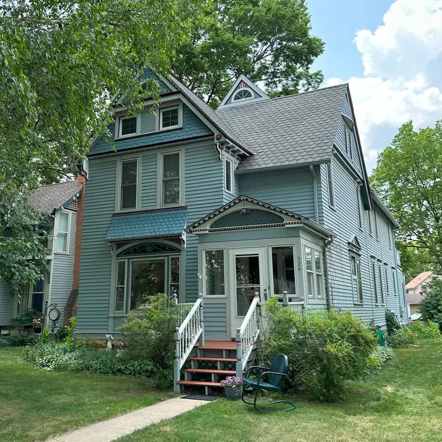 Three Story wood sided house with steep roof, painted in white, teal and burgundy. Enclosed Front Porch with 4 wood stairs and green metal rocker in yard.