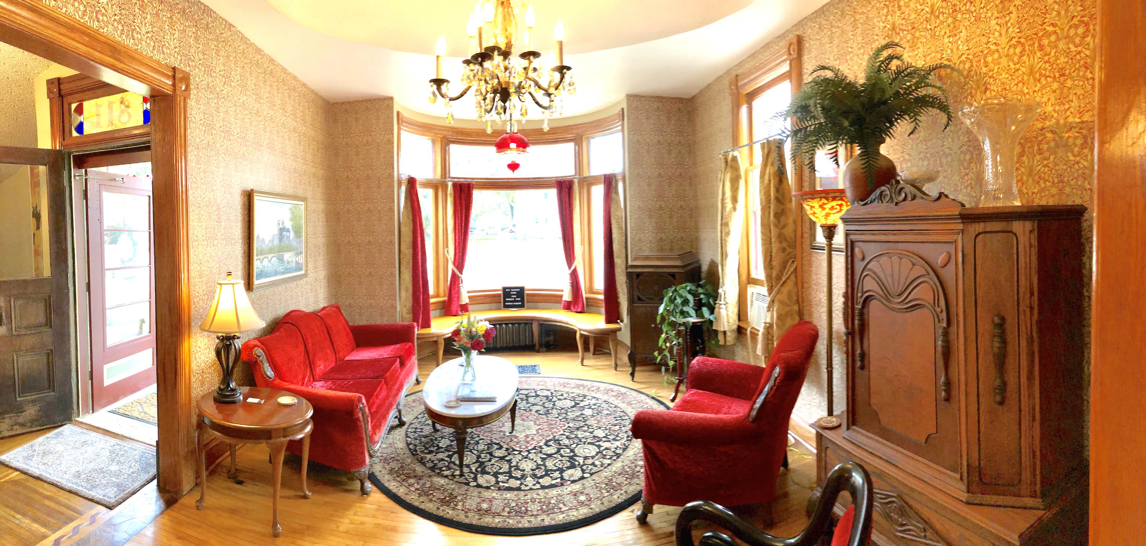 A vintage living room featuring red sofas, an ornate chandelier, and large windows with curtains.