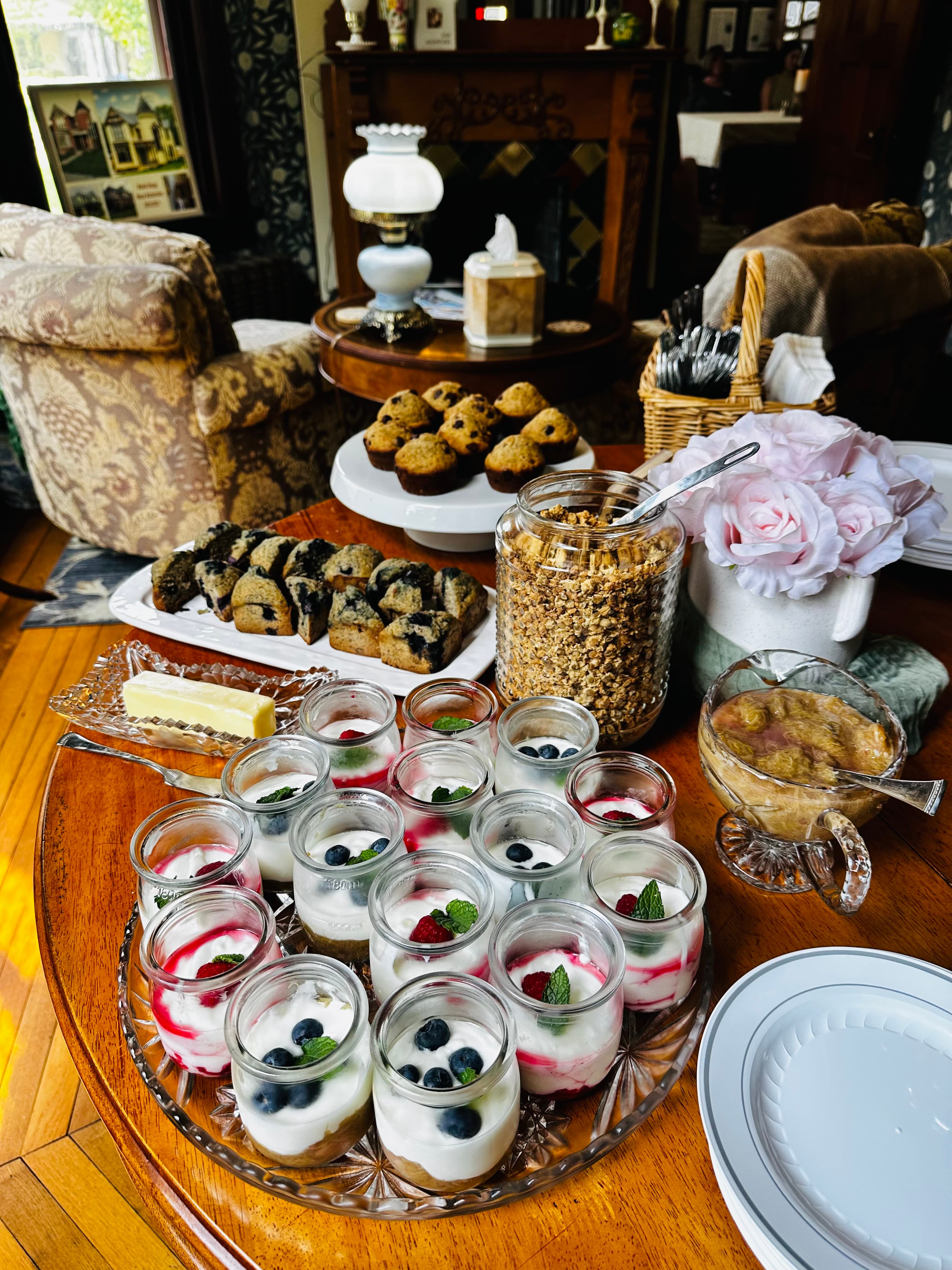 A beautifully arranged dessert table featuring assorted layered jars, muffins, granola, and delicate floral accents in a cozy setting.