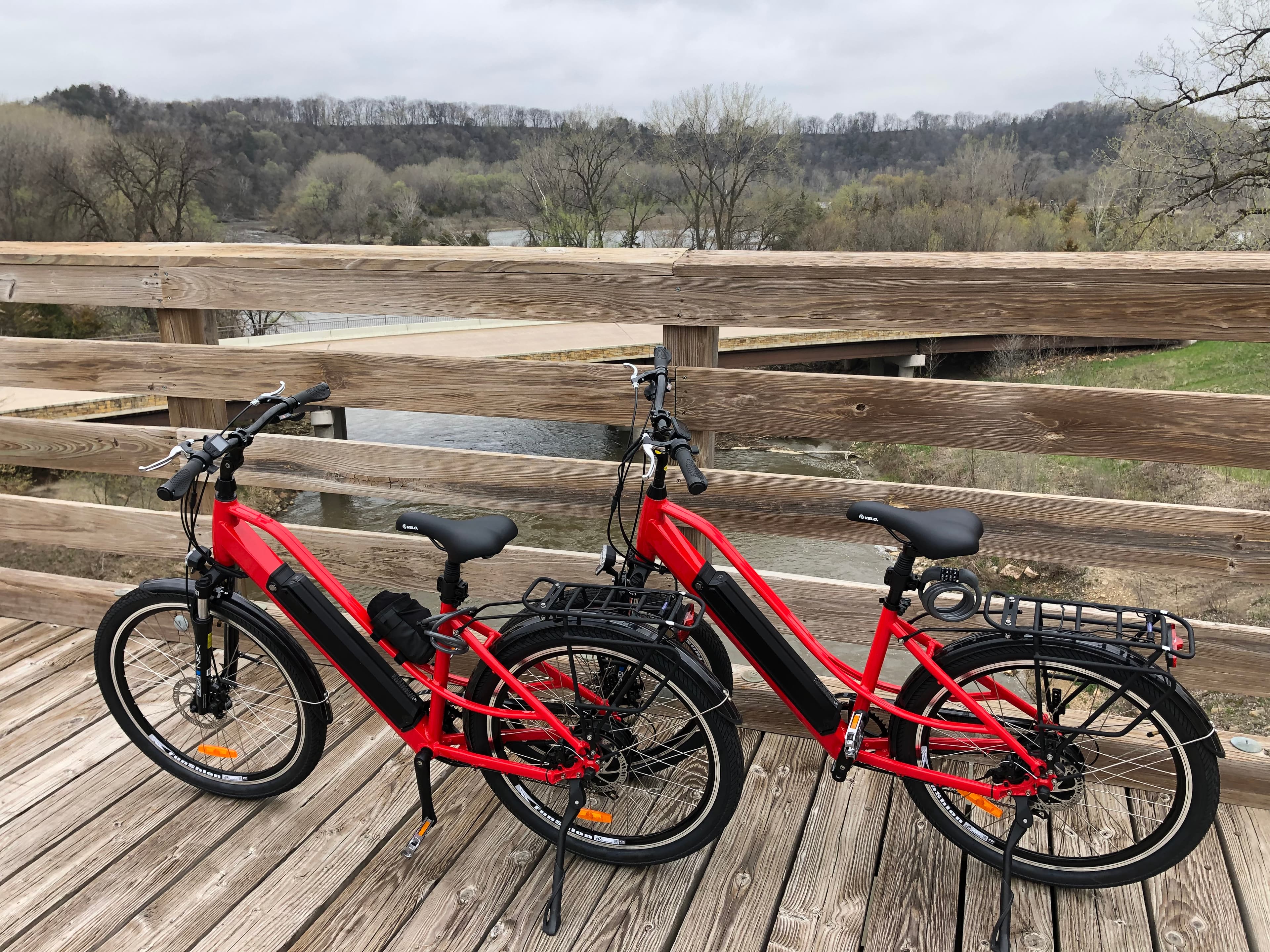 Two red electric bicycles parked on a wooden bridge overlooking a river and trees.