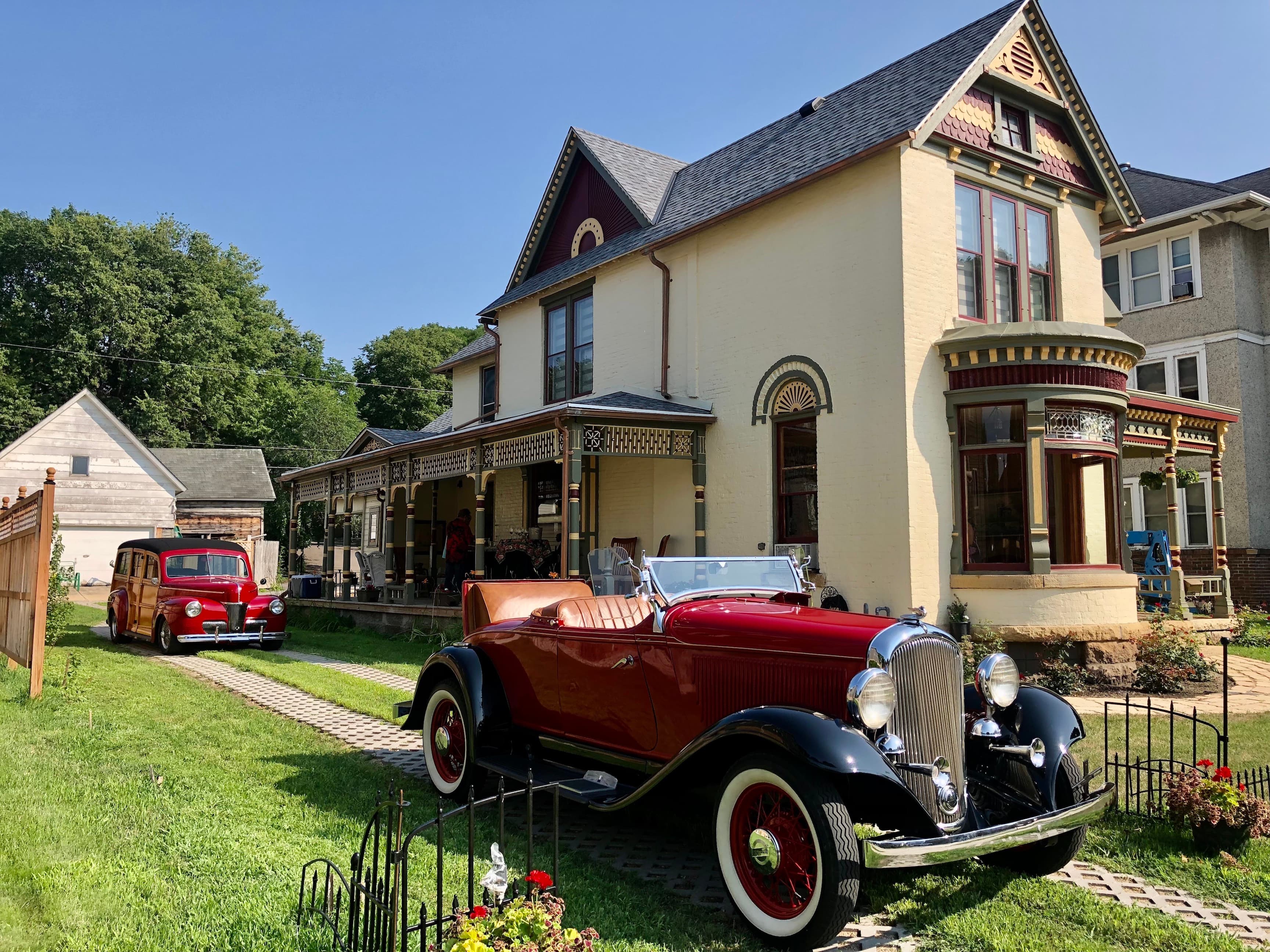 A red vintage car parked in front of a Victorian-style house on a sunny day.
