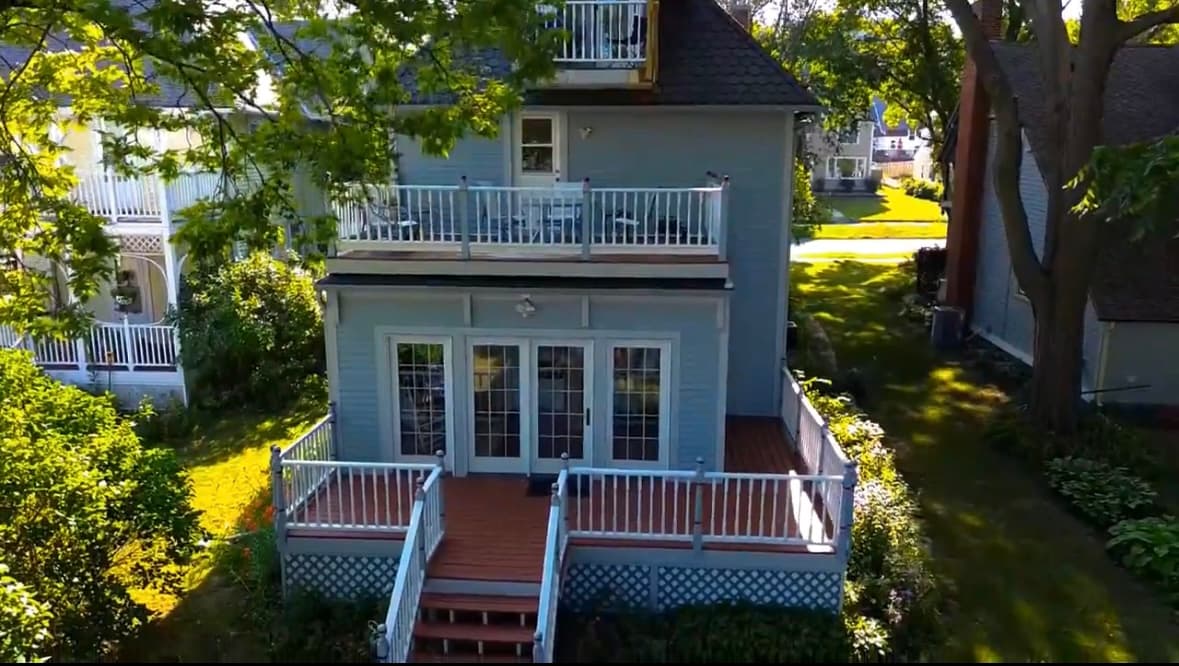 A two-story blue house with a wooden deck and a balcony surrounded by greenery.
