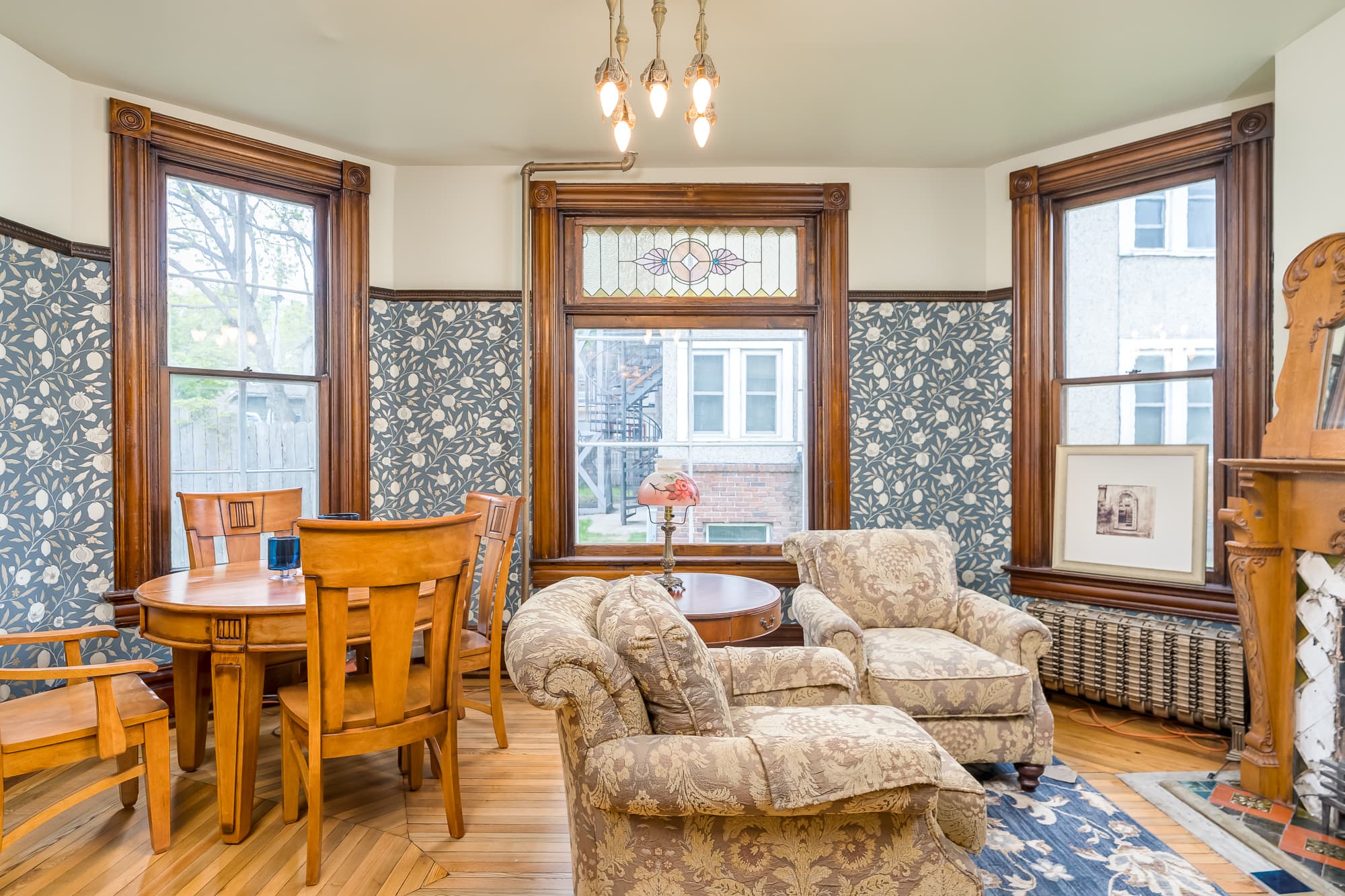 A cozy dining area featuring a wooden table, patterned chairs, and large windows with floral wallpaper.